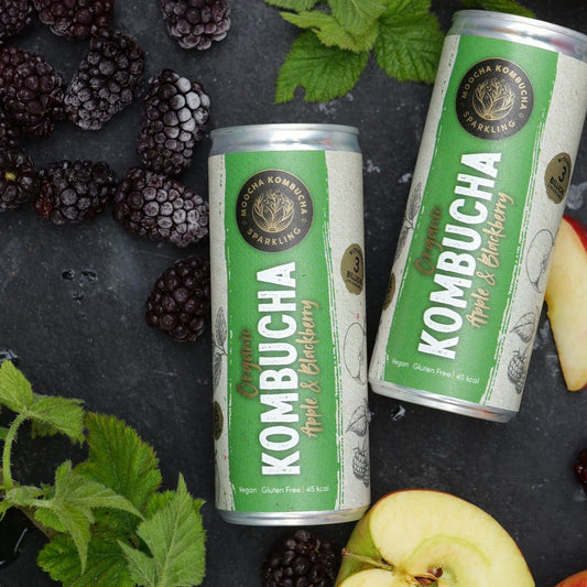 Two cans of Kombucha with blackberries and apples on a dark surface